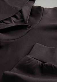 Primo piano di un indumento in tessuto scuro che mostra un colletto alto e un polsino arricciato su un capo a maniche lunghe.