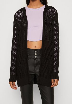 Femme portant un cardigan noir en maille ajourée sur un top court lavande et un pantalon noir taille haute avec trois boutons.