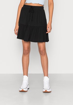 Jupe noire à volants en tissu doux, dotée d'une taille élastique avec un cordon de serrage. Le modèle porte des baskets blanches.