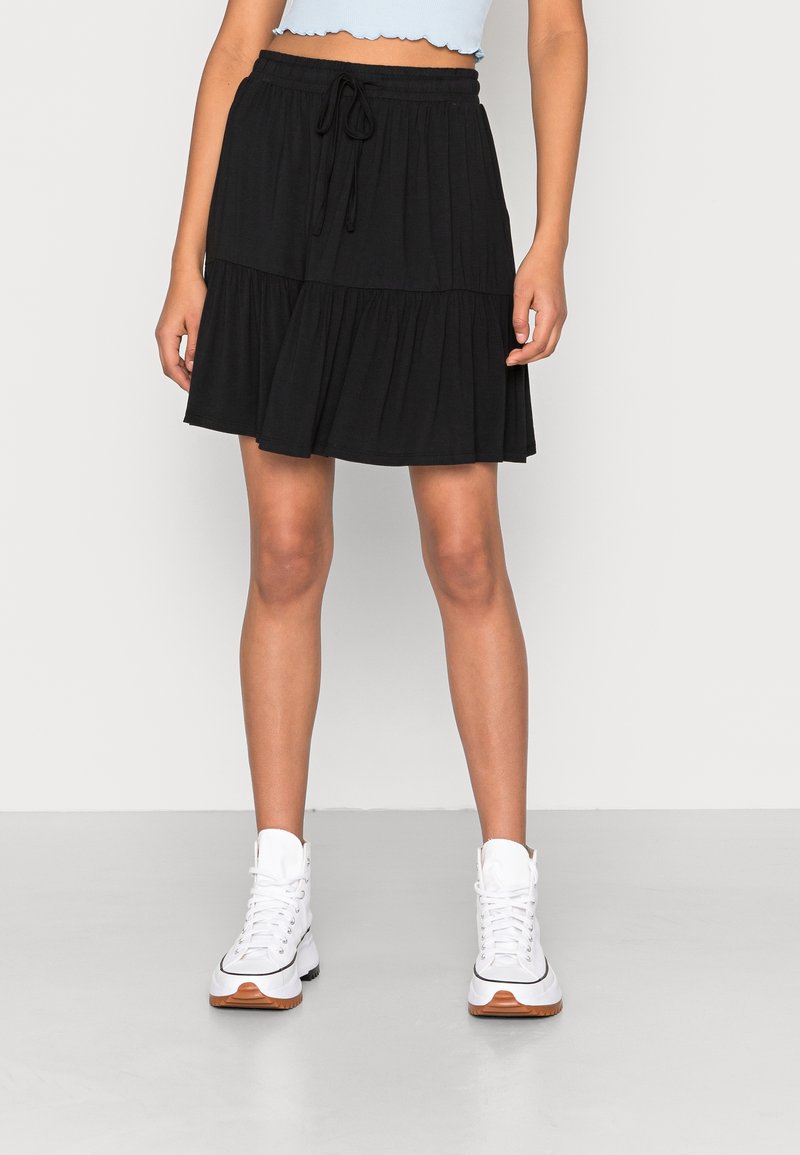 Jupe noire à volants en tissu doux, dotée d'une taille élastique avec un cordon de serrage. Le modèle porte des baskets blanches.