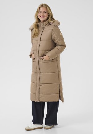 Geacă puffer lungă, bej, cu guler înalt, design cu panouri cusute, două buzunare frontale și închidere cu fermoar. Poartă peste blugi închisi la culoare și adidași.