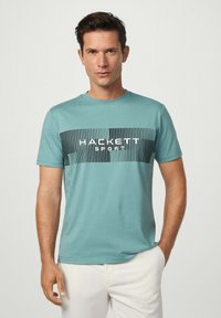 Muž v tyrkysovém tričku "Hackett Sport" a bílých kalhotách, stojící s jednou rukou v kapse na jednobarevném světlém pozadí.