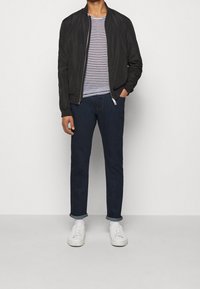 Osoba stoi na pierwszym planie, ubrana w czarną kurtkę bombową, w paski koszulę, ciemne dżinsy i białe trampki na neutralnym tle.