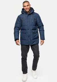 Marineblå pufferjakke med hætte, frontknapper, to store frontlommer og lynlåslommer på brystet. Tekstureret stof med kviltet design.