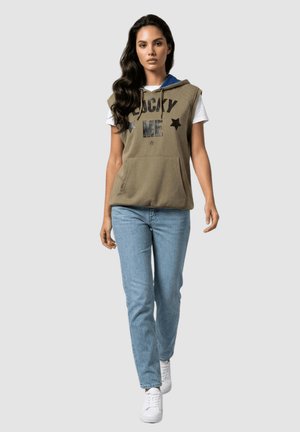 Mujer caminando con sudadera sin mangas con capucha en color caqui con el texto "Lucky Me", camiseta blanca, vaqueros azul claro y zapatillas blancas sobre fondo gris.
