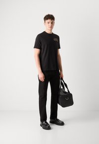 Sort t-shirt med et gult logo, parret med sorte jeans og sorte sko. Modellen holder en sort, struktureret duffel bag.