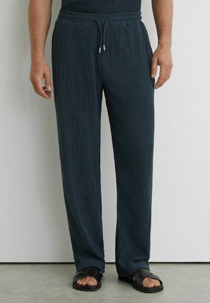 Mężczyzna w granatowych, teksturowanych spodniach z troczkami, noszący czarne sandały z odkrytymi palcami, stoi przy białej ścianie na szarej podłodze.