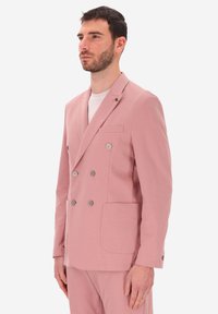 Uomo con capelli corti e scuri e barba, che indossa un blazer rosa doppiopetto e pantaloni abbinati, che guarda a sinistra su uno sfondo chiaro.