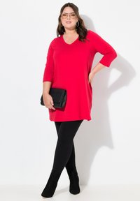 Rode tuniek met een V-hals, driekwart mouwen en een rechte pasvorm, gecombineerd met zwarte leggings en enkellaarsjes. Houdt een zwarte clutch vast.