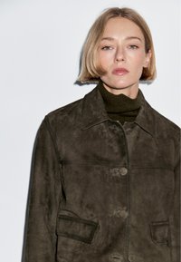 Tmavě olivově zelená semišová bunda s špičatým límcem, zapínáním na knoflíky vpředu a dvěma náprsnými kapsami. Hladká textura s jemnými šicími detaily.