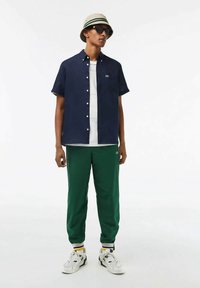 Navy short-sleeve button-up shirt, white T-shirt underneath, green track pants with striped cuffs, beige bucket hat, black sunglasses, white sneakers.