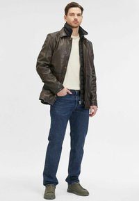 Braune Lederjacke mit schwarzem wattiertem Innenfutter, getragen über einem cremefarbenen Hemd, kombiniert mit blauen Jeans und olivgrünen Sneakern.