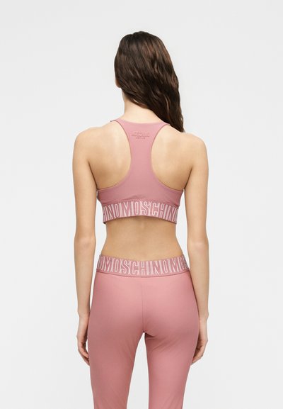 Γυναίκα με μακριά καστανά μαλλιά, φορώντας ροζ αθλητικό σουτιέν και κολάν με λωρίδες λογότυπου "MOSCHINO", εμφανιζόμενη από πίσω.