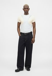 Homme aux cheveux blonds courts portant un haut blanc moulant avec des panneaux beige aux épaules, pantalon large noir, ceinture noire et chaussures noires, debout.