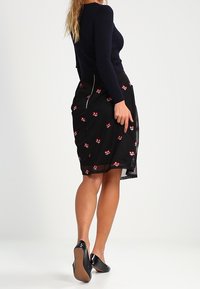 Gonna nera con ricami floreali rossi, sovrapposizione semi-trasparente e zip laterale. Abbinata a un top aderente a coste blu navy e tacchi neri.