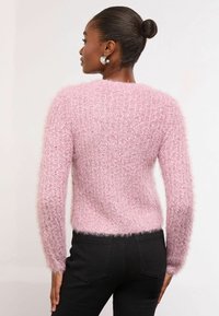 Rozā pūkains džemperis, kas izgatavots no teksturēta materiāla, ar garām piedurknēm un īsu dizainu. Kombinēts ar melniem augsto jostasvietu džinsiem.