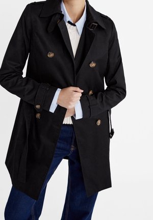 Person wearing a black double-breasted trench coat over a white sweater and blue collared shirt, paired with blue jeans.