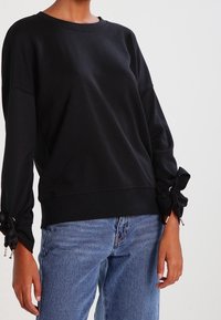 Sudadera negra de manga larga con cuello redondo, que presenta detalles con lazo en las muñecas y un corte relajado, combinada con jeans de color azul claro.