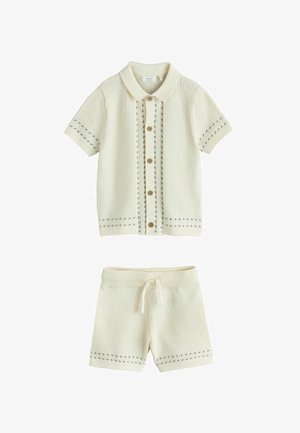 Kremasta pletena košulja kratkih rukava s kopčanjem na tkanice i ovratnikom, te odgovarajuće šorc s vezicama, oba s diskretnim sivim uzorkom šavova.