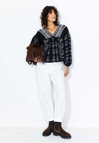 Blouse noire brodée avec un col en V et des manches longues, assortie à un pantalon blanc à coupe décontractée et des bottines en daim marron.