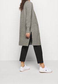 Person i en lång grå stickad kofta med sidofickor, svarta cropped byxor och vita sneakers som går mot en enkel vit bakgrund.