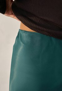 Teal satin nederdel med en glat tekstur, parret med en mørk ribbet sweater ved taljen. Beklædningen har en tætsiddende silhuet med glans.