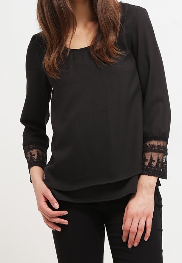 Femme portant une blouse noire à manches longues avec des manches en dentelle transparente, associée à un pantalon noir, debout devant un fond blanc.
