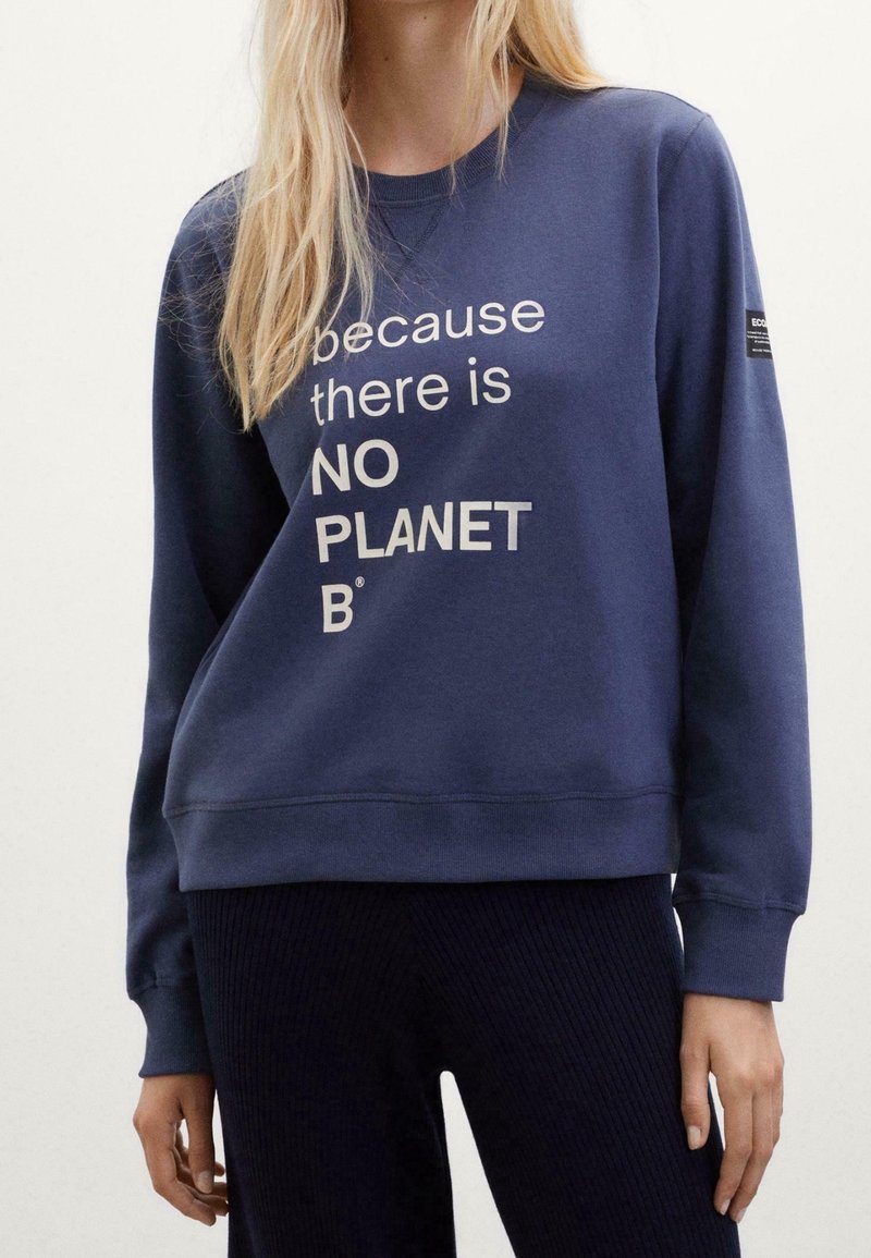 Mujer vestida con un sudadera azul marino con el texto "porque no hay UN PLANETA B" y pantalones oscuros de canalé, de pie contra un fondo claro.