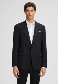 Giacca da abito nera su misura con rever a destra, due bottoni e tasche a pattina. Abbinata a una camicia bianca con colletto discreto.