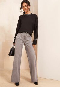 Maglione nero a maglia con dettagli argentati, abbinato a jeans grigi a vita alta e a gamba larga. Il modello tiene in mano una piccola borsa nera e indossa scarpe con tacco a punta.
