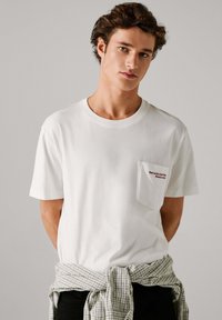 Camiseta blanca de manga corta con diseño liso, que presenta un bolsillo en el pecho izquierdo con texto bordado. Se lleva con una franela de cuadros gris atada a la cintura.