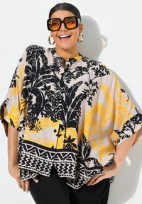 Blouse en soie beige avec des motifs floraux noirs et jaunes, dotée d'un col haut, d'une fermeture à boutons et de larges manches avec des accents géométriques.