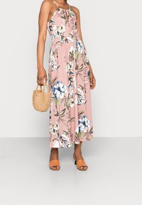 Femme portant une robe midi sans manches à fleurs roses, tenant un petit sac tissé, et portant des talons ouverts couleur beige.