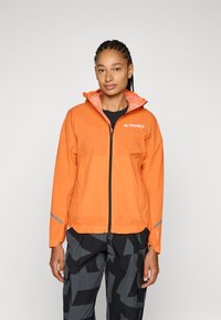 Orangen wasserdichte Jacke mit Reißverschluss vorne, verstellbarer Kapuze und reflektierenden Akzenten. Kombiniert mit dunkel gemusterten Cargo-Hosen.