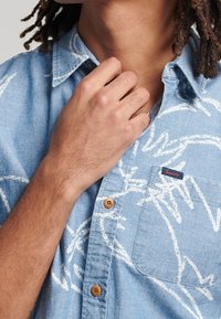 Hellblaues, kurzärmeliges Jeanshemd mit weißen abstrakten Mustern, einer Brusttasche und braunen Knöpfen.