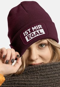 Burgundy knit beanie with a rectangular white patch reading "IST MIR EGAL." Features a ribbed texture and snug fit.