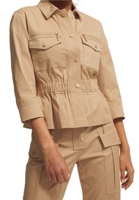 Mujer vestida con una chaqueta utilitaria beige con botones de presión en el frente y bolsillos con cremallera, combinada con pantalones cargo a juego.
