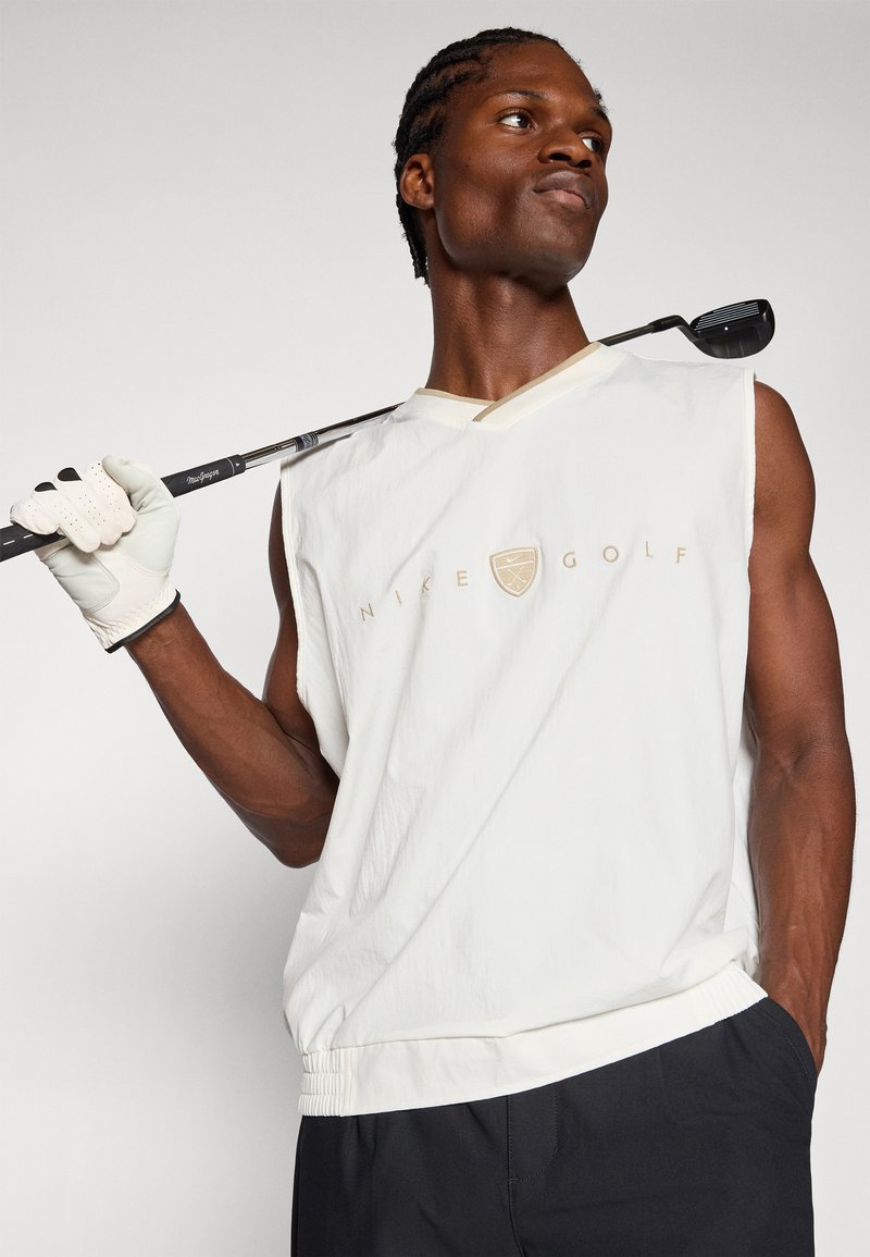 Man wearing white sleeveless Nike Golf top and black pants, holding golf club over shoulder, looking to the side.