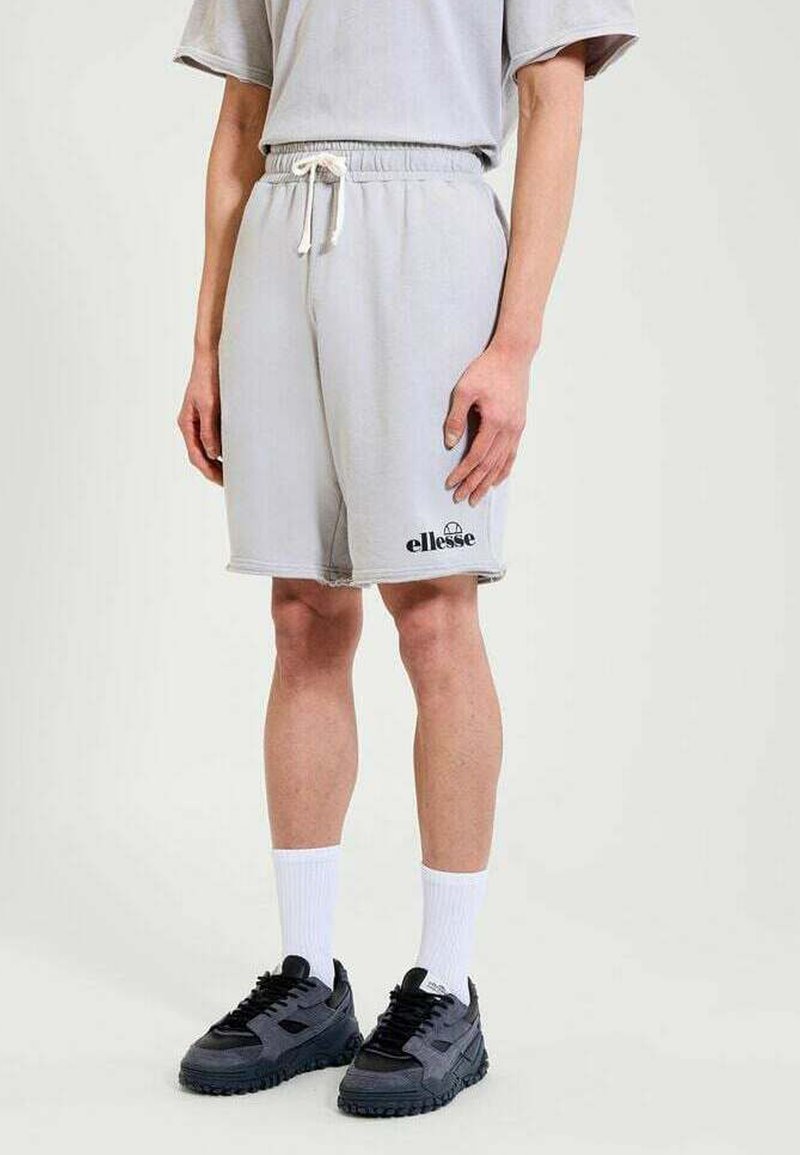 Persona vistiendo pantalones cortos gris claro de ellesse con cordón blanco, calcetines deportivos blancos y zapatillas deportivas negras, de pie sobre un fondo liso.