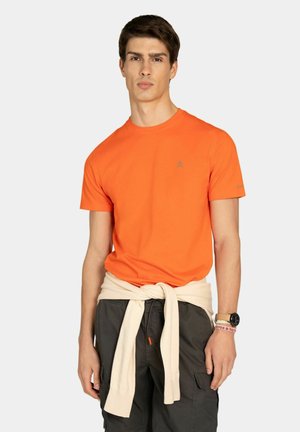 Joven con camiseta naranja brillante y pantalones cargo oscuros, con un suéter beige atado alrededor de la cintura, sobre un fondo liso.