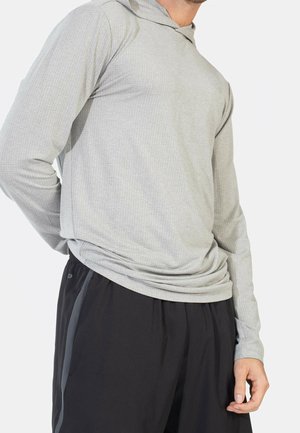 Mężczyzna w jasnoszarej teksturowanej bluzie z kapturem z długim rękawem i czarnych spodniach sportowych z ciemnymi paskami po bokach na białym tle.