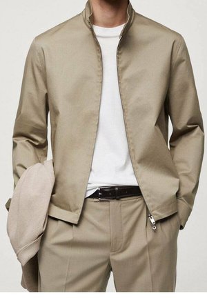 Mann trägt beige Reißverschlussjacke über weißem T-Shirt und passenden beigen Hosen, hält einen beigen strukturierten Stoff in der linken Hand.