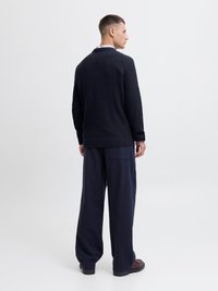 Marineblauer, strukturiertes Strickpullover mit Schalkragen, kombiniert mit weit geschnittenen marineblauen Hosen und braunen Schuhen, von hinten betrachtet.