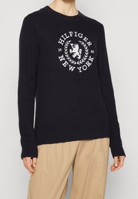 Marineblauer Rippenpullover mit einem weißen gestickten Logo "Hilfiger New York" und einem Löwen innerhalb eines Lorbeerkranzes, rundem Ausschnitt und langen Ärmeln.