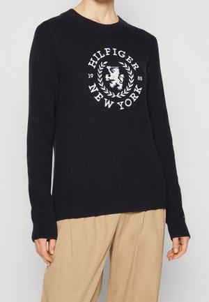 Granatowy sweter ze ściągaczem z białym haftowanym logo "Hilfiger New York" oraz lwem w laurowym wieńcu, okrągłym dekoltem i długimi rękawami.