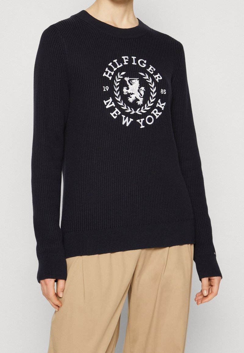 Marineblauer Rippenpullover mit einem weißen gestickten Logo "Hilfiger New York" und einem Löwen innerhalb eines Lorbeerkranzes, rundem Ausschnitt und langen Ärmeln.