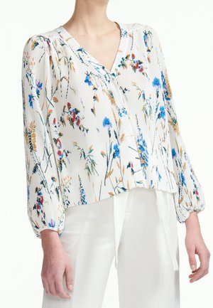 Femme portant un chemisier blanc à manches longues avec un motif floral multicolore et un pantalon blanc taille haute.