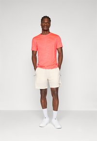 Camiseta de manga corta color coral con un patrón sutil, combinada con pantalones cortos beige. Los accesorios incluyen calcetines blancos y zapatillas deportivas. Look simple y deportivo.