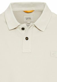Beige polo skjorte med tekstureret overflade, to sorte knapper ved kraven og et logo på venstre bryst. Etiketten angiver størrelse L.