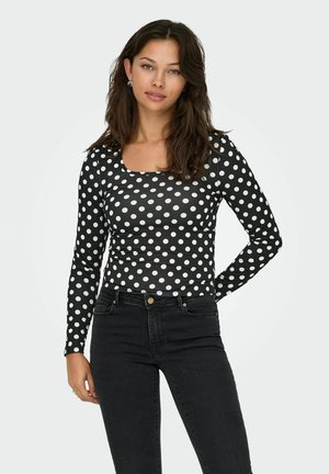 Top nero a maniche lunghe con motivo a pois bianchi e scollatura quadrata, abbinato a jeans neri. Tessuto liscio e design aderente.